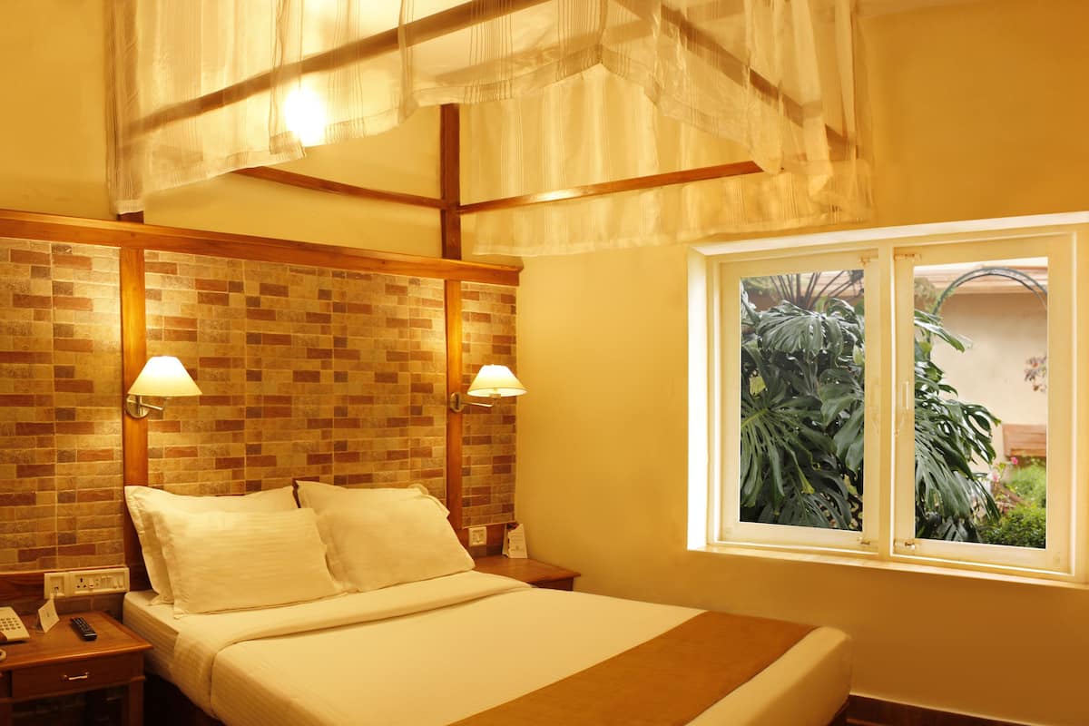 Habitación de hotel con pared de ladrillo decorativa, cama con ropa de cama blanca, mesita de noche con lámpara, escritorio con un jarrón de flores y ventana con vistas a zonas verdes.