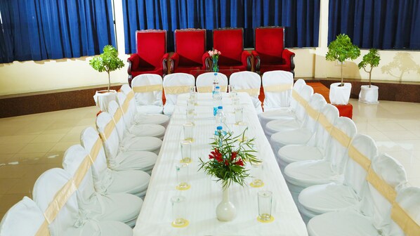 Une salle de banquet avec une longue table dressée pour un événement formel, des chaises rouges en arrière-plan et des plantes en pot de chaque côté.