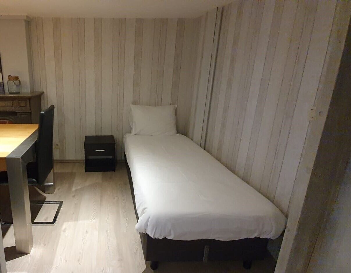 Een kleine kamer met een enkel bed, een houten bureau en stoel, een nachtkastje en een klein dressoir.