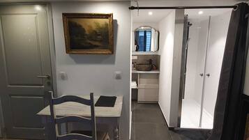 A narrow hallway with a door leading to a room with a table and chair, a painting on the wall, and a bathroom with a shower and sink.