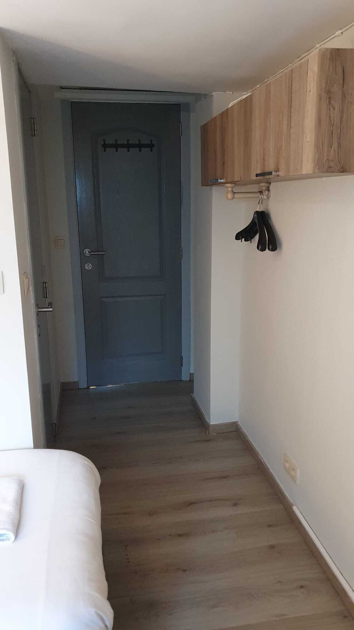 A narrow hallway with a wooden door at the end, a white wall with a wooden shelf above, and a bed with a white cover on the left.