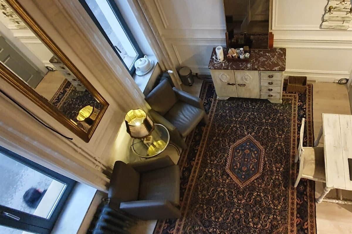 Una habitación con un ventanal grande, alfombra estampada, sofá, mesita de noche con lámpara y cómoda con cajones.