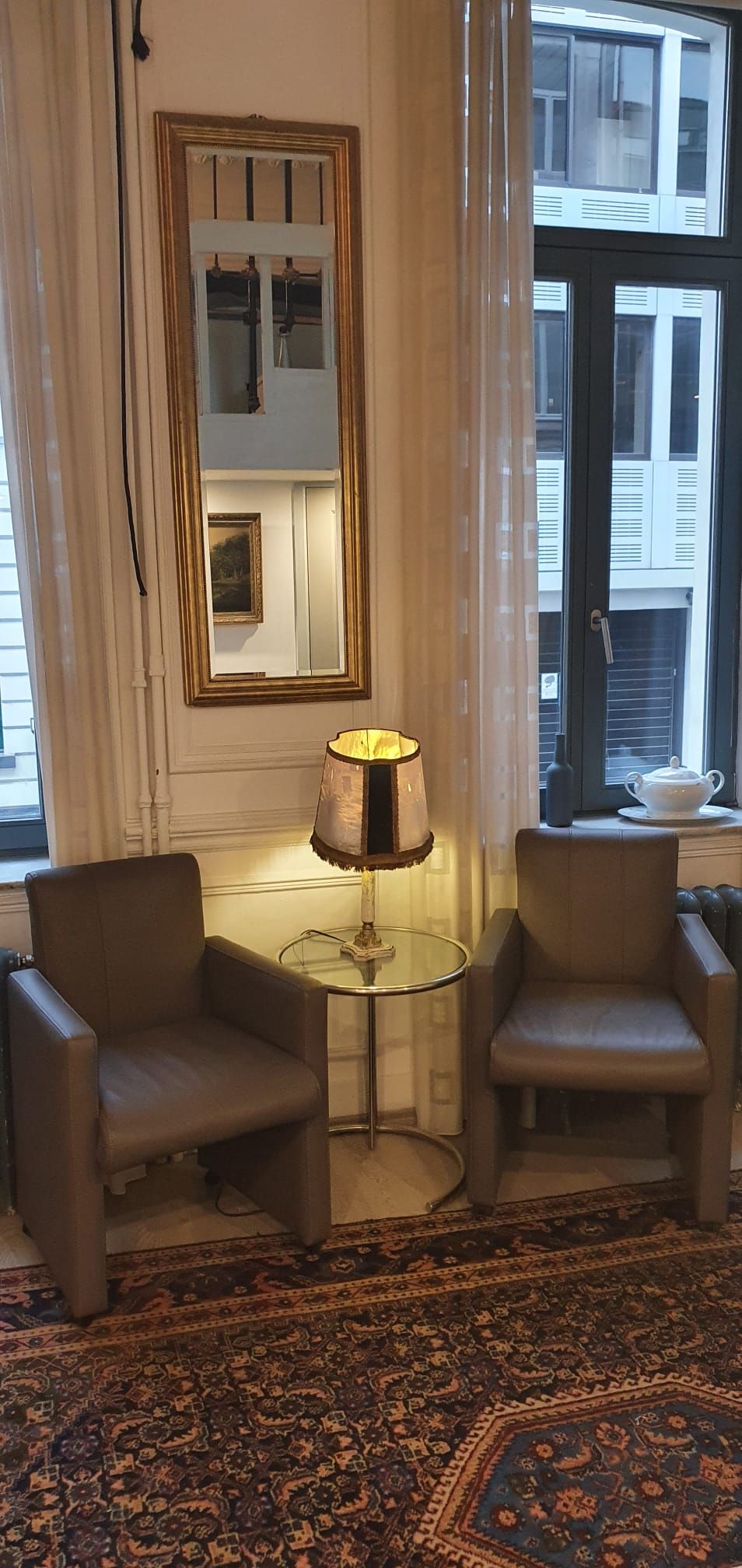 A room with two armchairs, a small table, a lamp, a patterned rug, and a window with blinds.