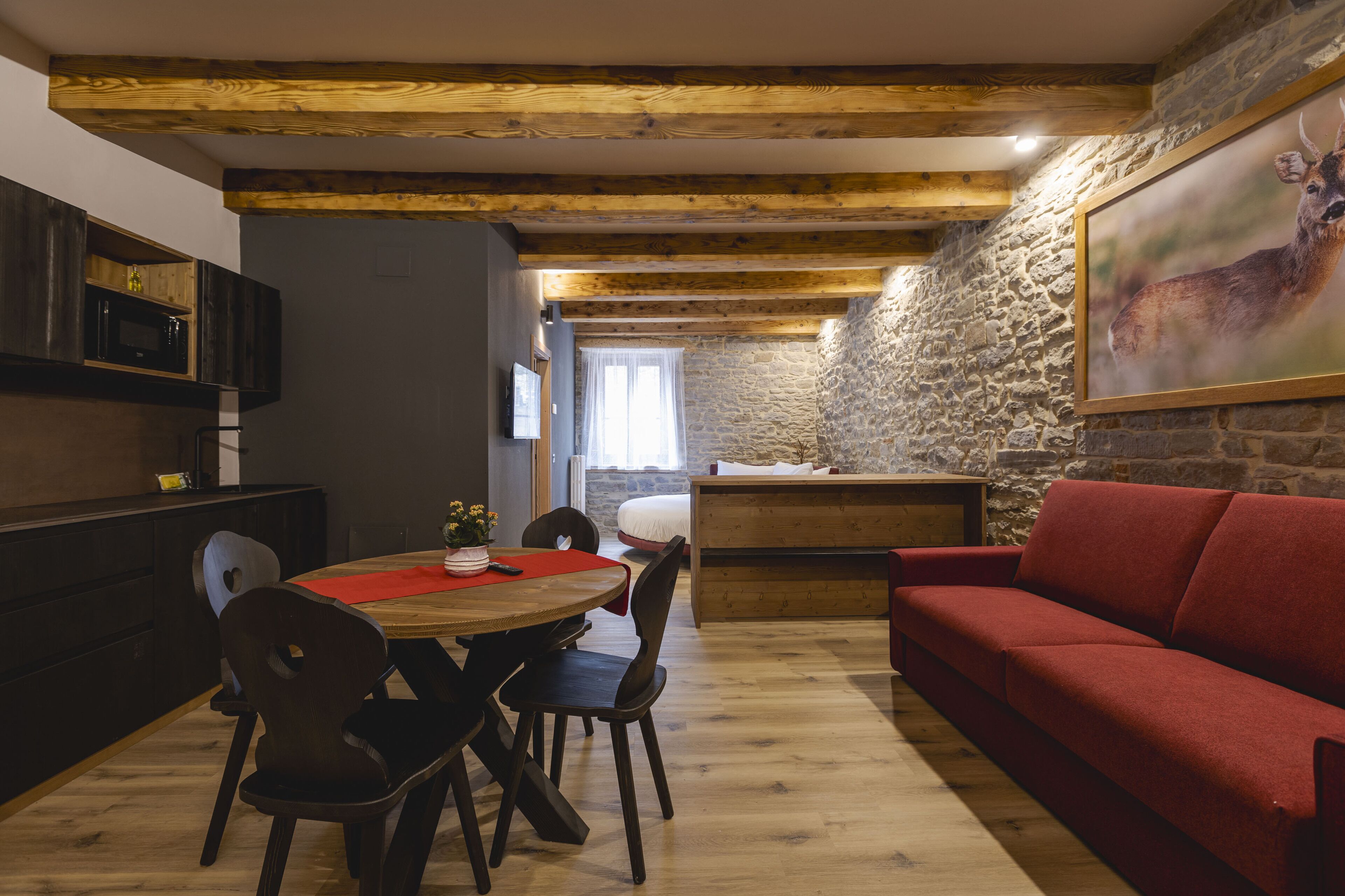 A cozy dining area with a wooden table and chairs, a red sofa, and a stone wall with a deer painting.