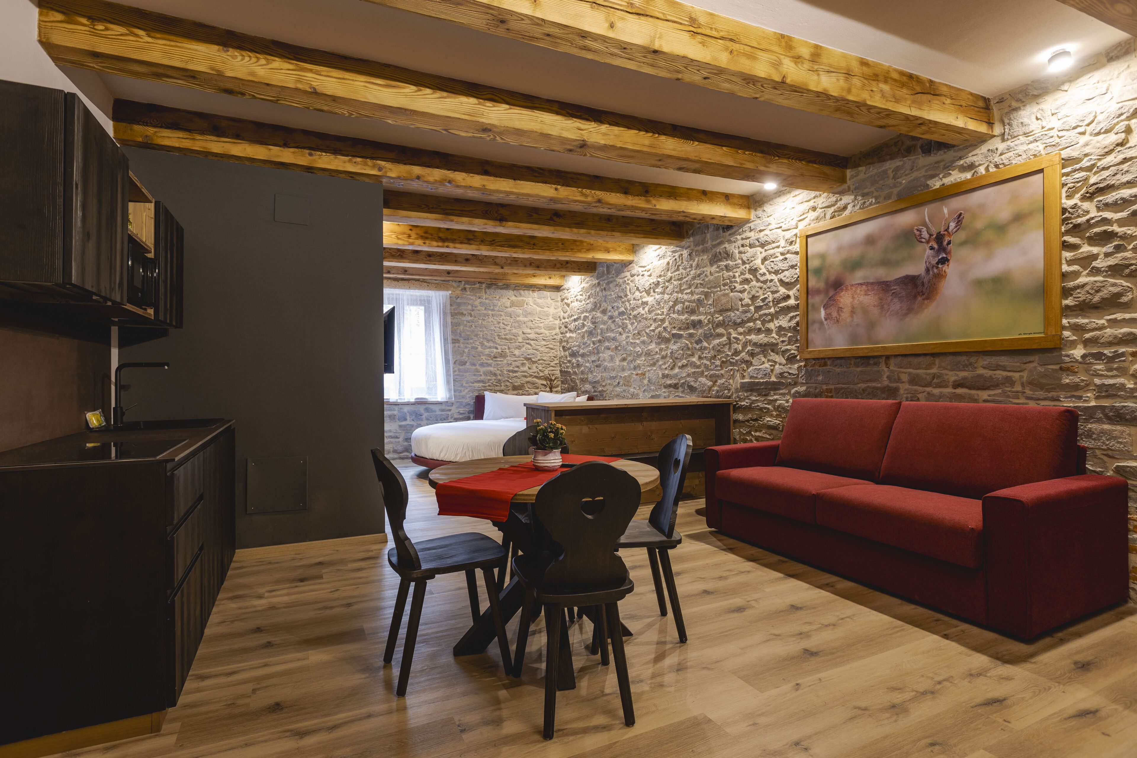 Ein gemütlicher Wohnbereich mit einem roten Sofa, einem Holzesstisch und Stühlen. Ein Bild von einem Reh hängt an der Wand.
