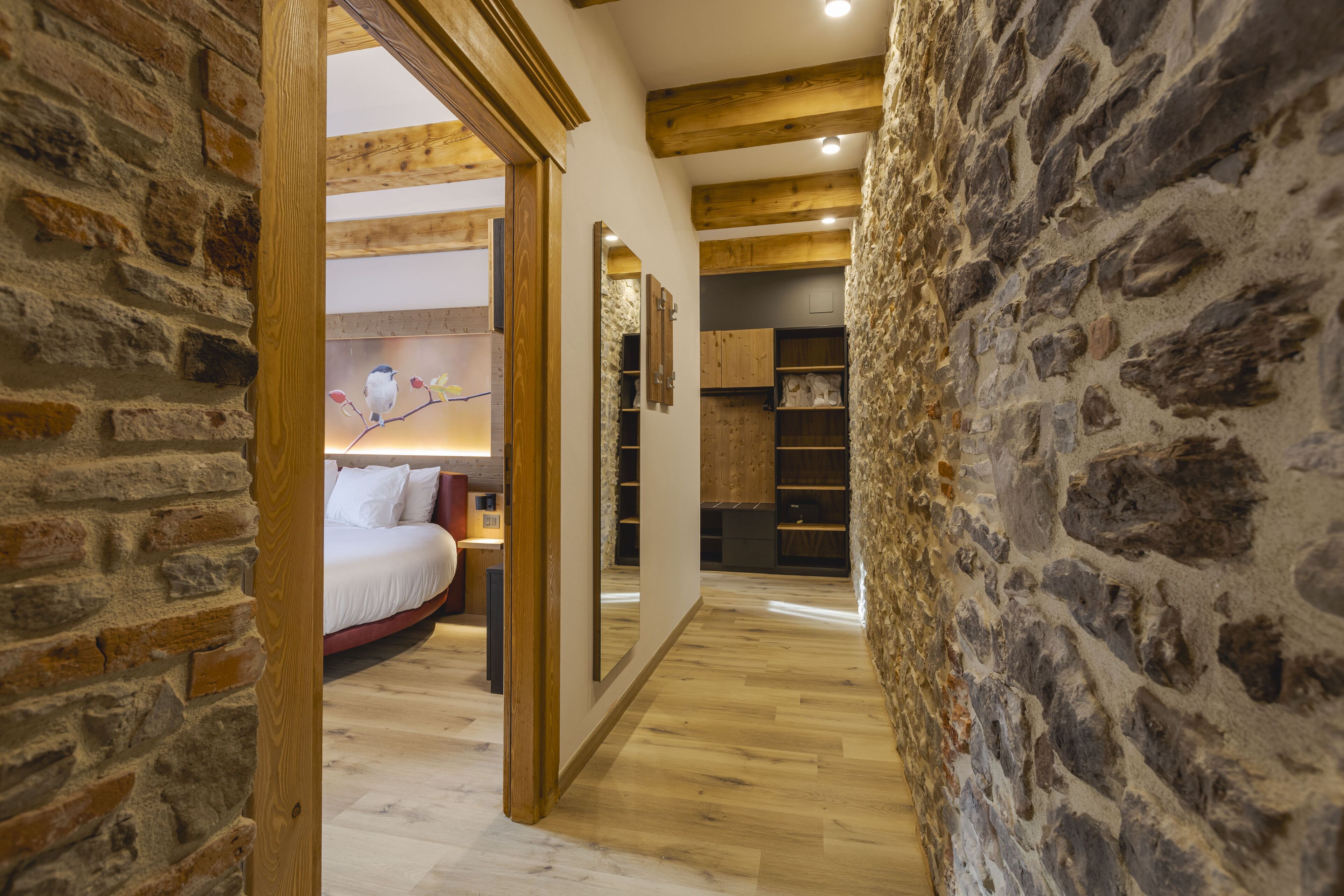 A narrow hallway with stone walls and wooden beams, leading to a bedroom with a bed and a nightstand.