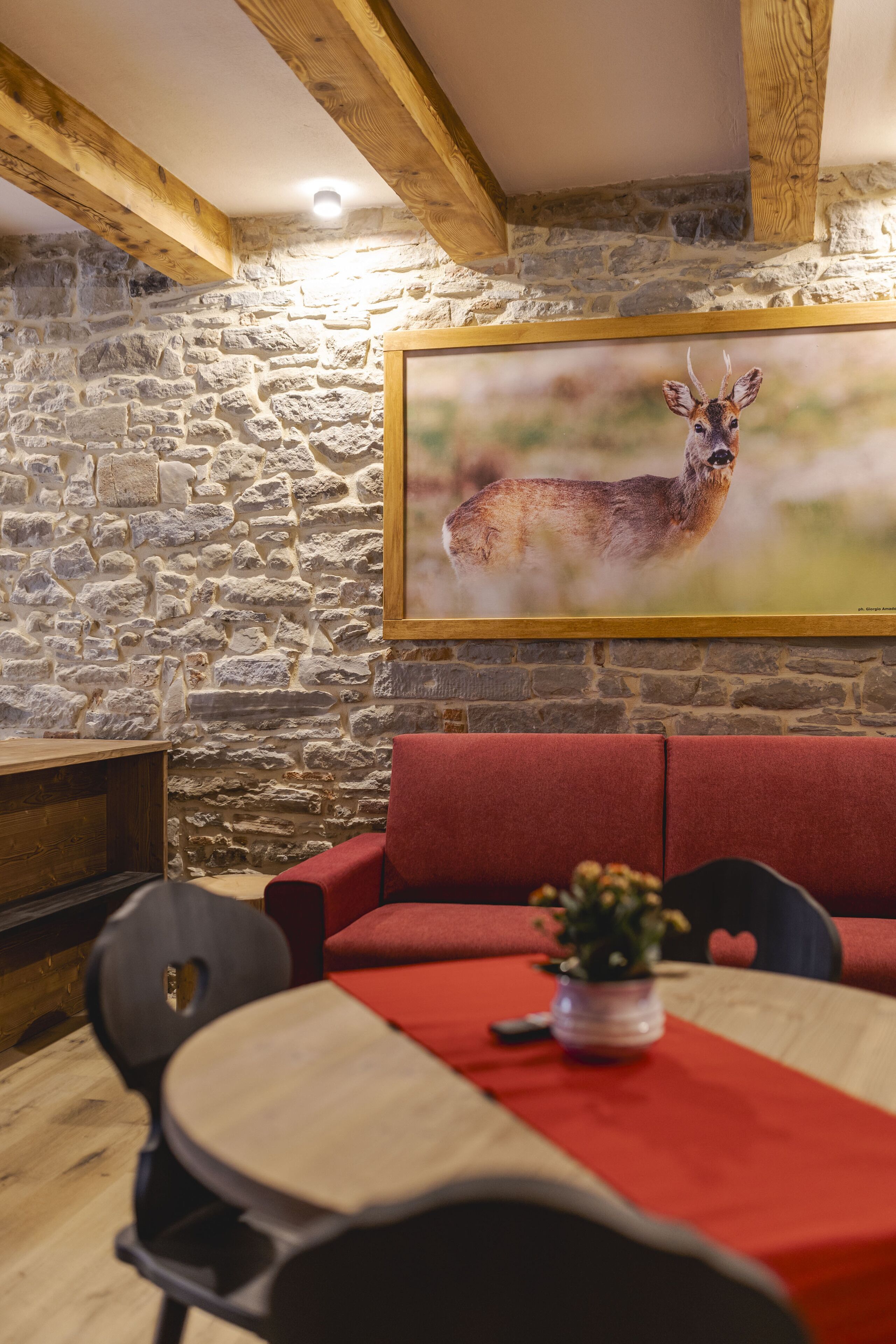 Una habitación rústica con paredes de piedra, techo de madera, un sofá rojo, una mesa de comedor con sillas y un cuadro de un ciervo enmarco.
