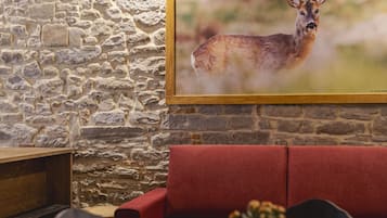 Una habitación rústica con paredes de piedra, techo de madera, un sofá rojo, una mesa de comedor con sillas y un cuadro de un ciervo enmarco.