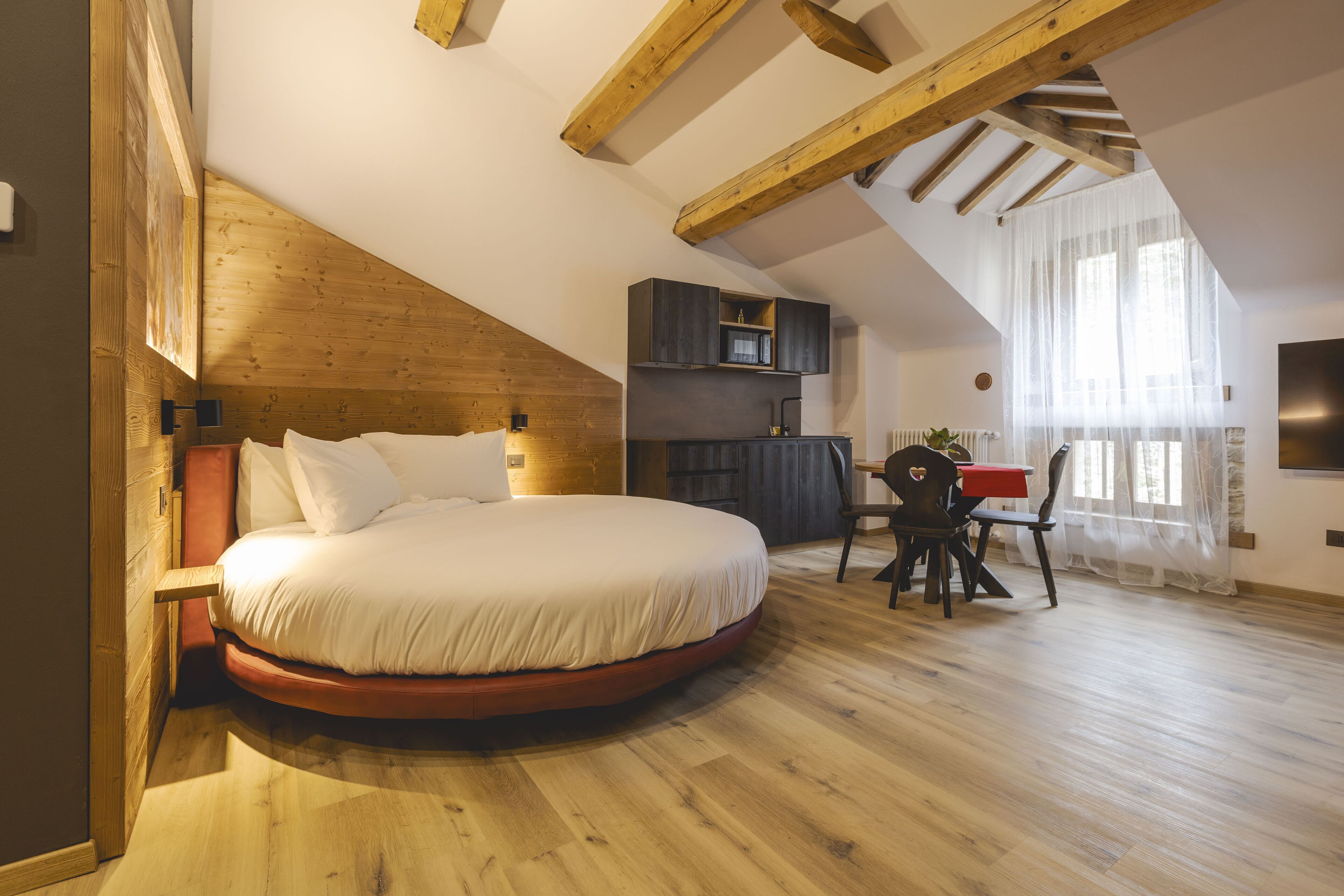 Ein Schlafzimmer mit einem runden Bett, Holzbalkendecke, einer kleinen Küchenzeile und einer Essecke mit Tisch und Stühlen.