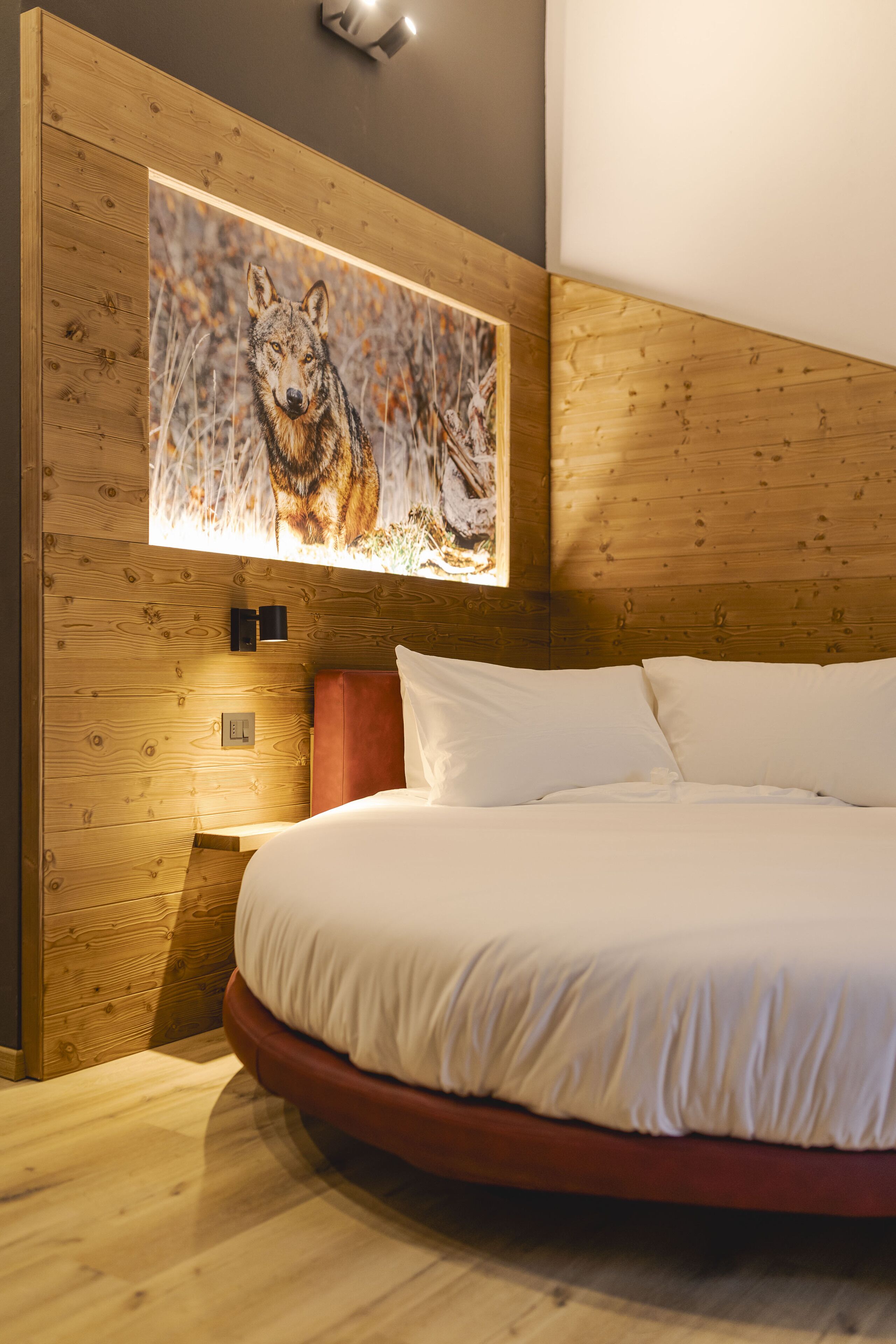 Ein Schlafzimmer mit Holz-Kopfteil, einem großen Gemälde eines Wolfs, einer roten Akzentwand und einem runden Bett mit weißer Bettwäsche.