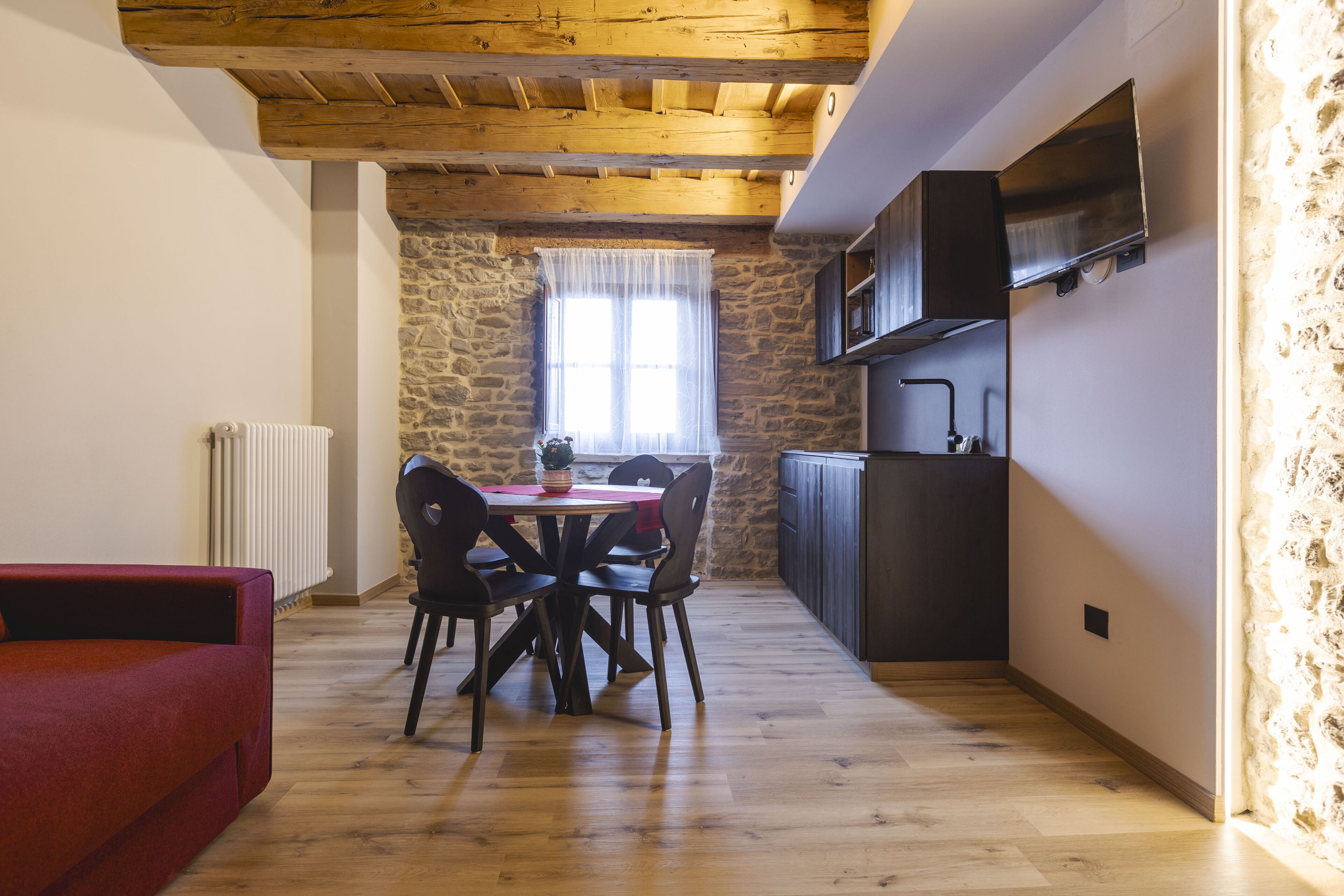 Une cuisine chaleureuse avec un mur en pierre, des poutres en bois au plafond, une table à manger avec des chaises et un canapé rouge.