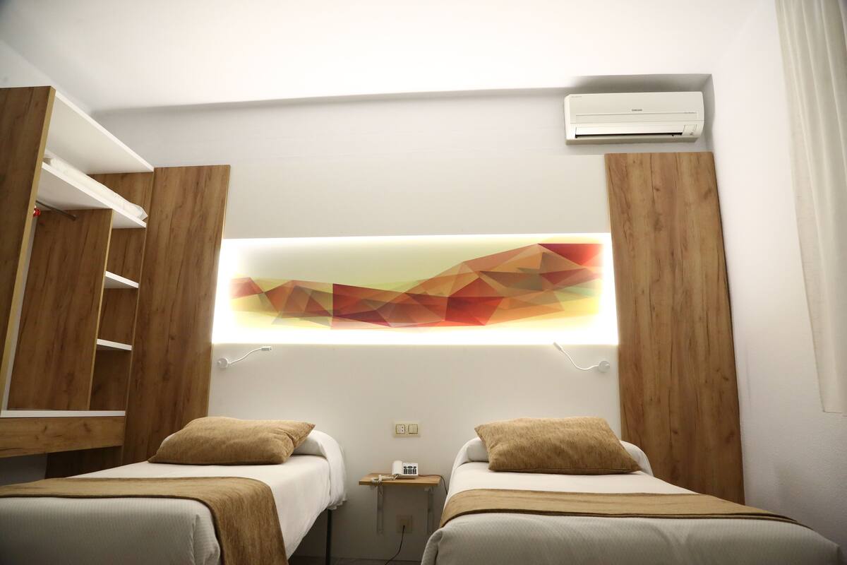 A hotel room with two beds, a wall-mounted air conditioner, and a geometric wall art piece.