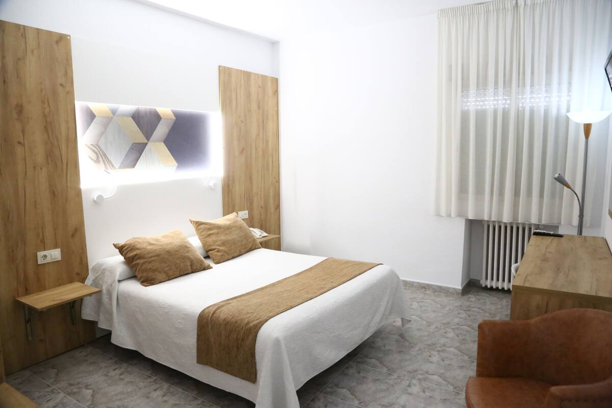 A hotel room with a bed, wooden headboard, a small wooden side table, a chair, and a lamp.