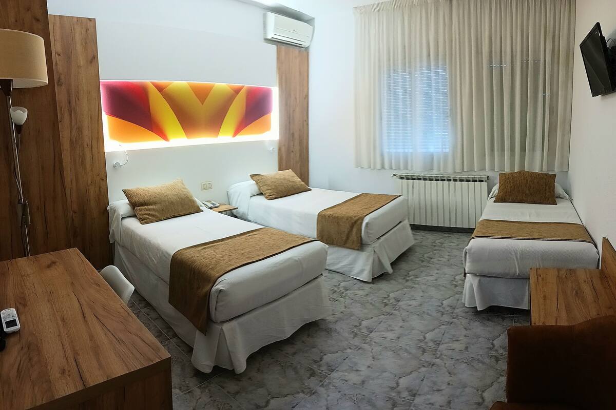 A hotel room with two single beds, a wooden desk, a lamp, a wall-mounted TV, and a radiator.