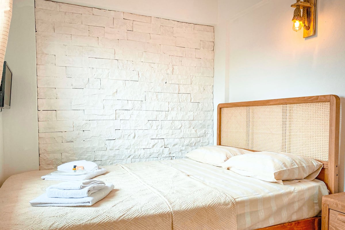 Una cama bien hecha con un cabecero tejido, una pared con diseño de ladrillo y una lámpara fijada a la pared.