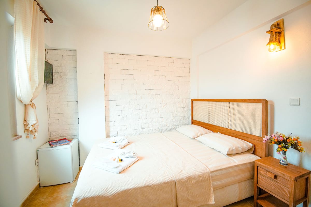 Una cama bien hecha con cabecera de madera, una mesita de noche con un florero y una pequeña mesita blanca con una revista.