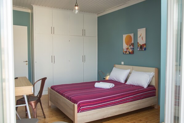 A bedroom with a bed, a desk, and a chair. There is a wardrobe and artwork on the wall.