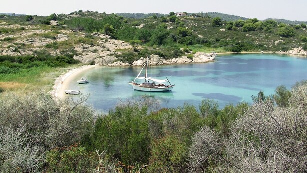 A coastal scene with a boat anchored near a sandy beach surrounded by rocky terrain and dense greenery. - Ariadne Eco Retreat (Polygyros)