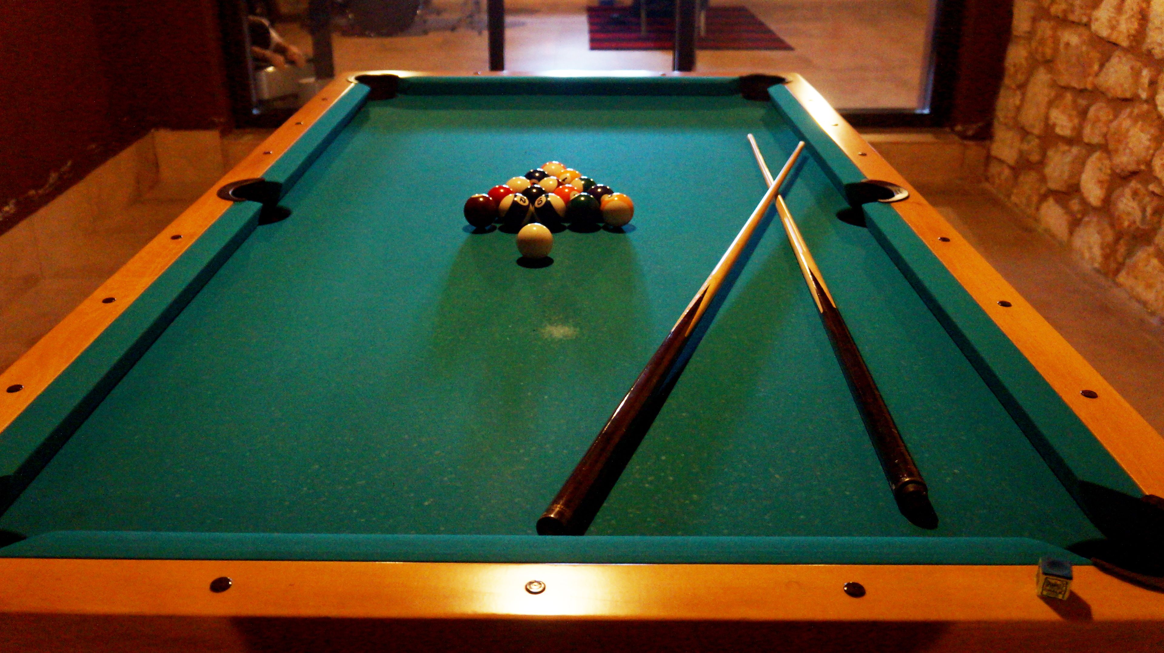 A pool table with balls and cues, surrounded by a stone wall and a wooden floor.