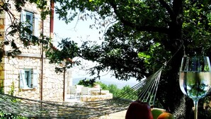 A hammock, a book, glasses, and fruit on a table with a view of a stone building and trees. - Ariadne Eco Retreat (Polygyros)