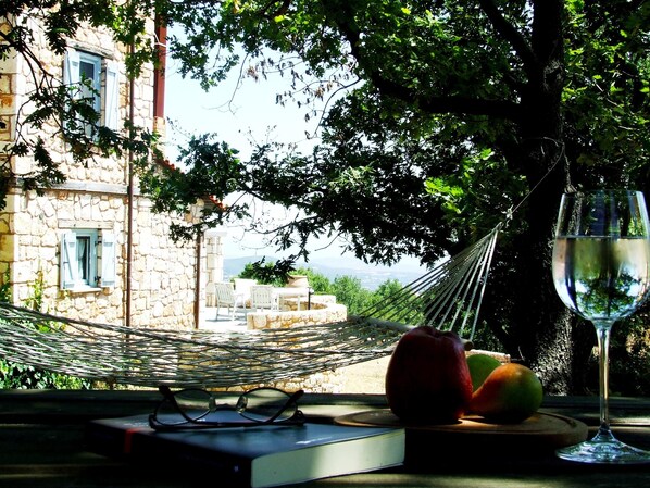 Eine Hängematte, ein Buch, eine Brille und Obst auf einem Tisch mit Blick auf ein Steinhaus und Bäume.