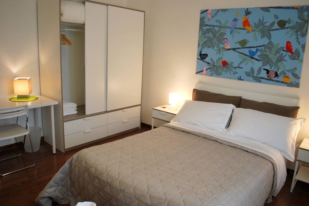 A hotel room with a bed, a desk, a chair, a wardrobe, and a painting on the wall.