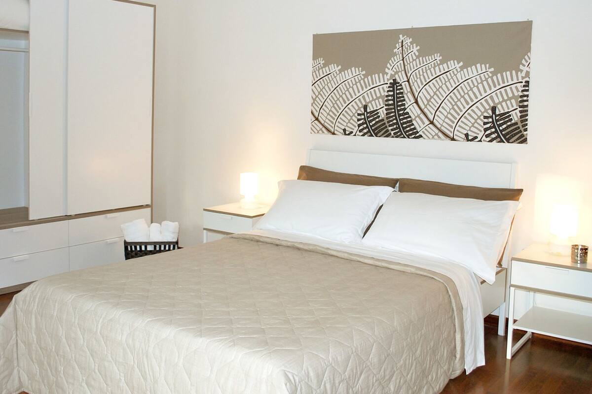 A neatly arranged hotel room with a large bed, two bedside tables, a nightstand, a mirror, and a wall-mounted artwork.