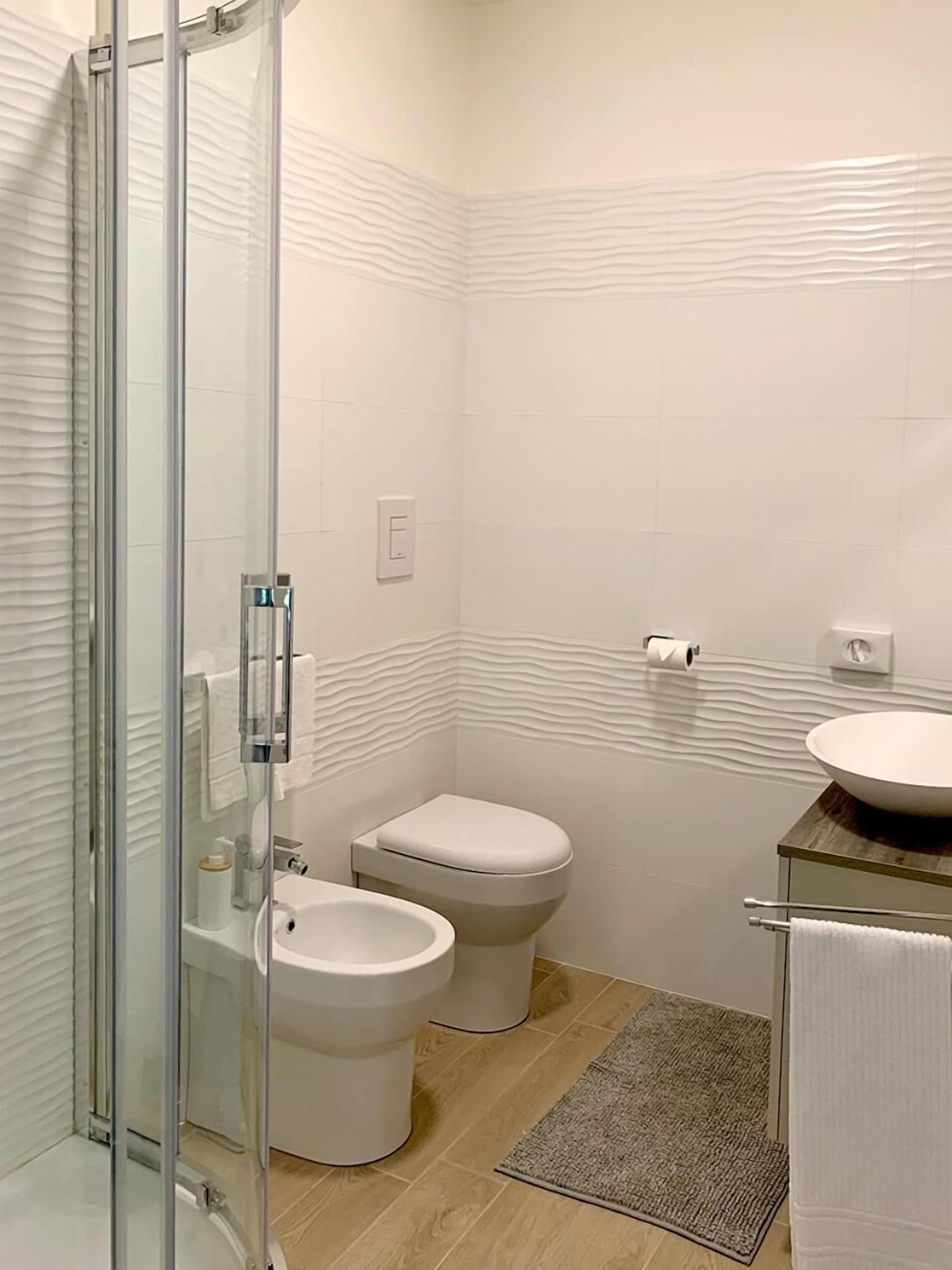 A modern bathroom with a glass shower enclosure, a toilet, a bidet, and a sink with a wooden countertop.