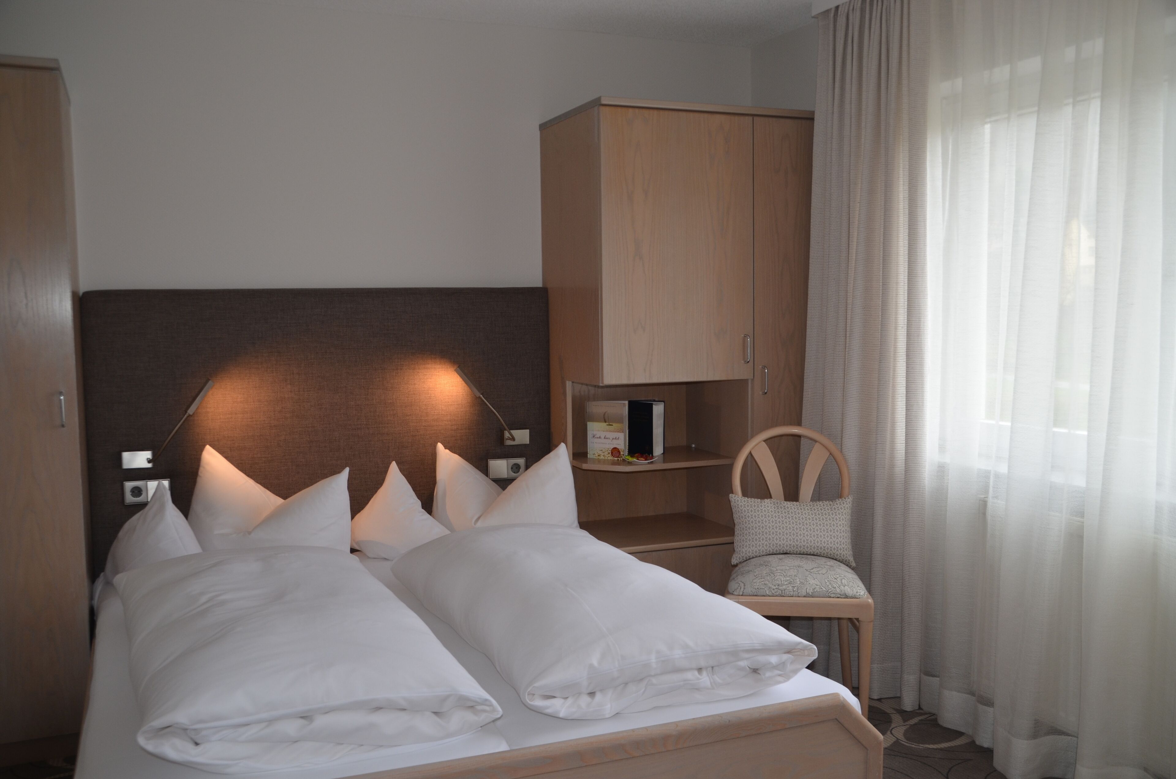 Ein ordentlich bezogenes Bett mit weißen Leinentüchern und Kissen, ein Holz-Kopfstück, ein Nachttisch mit Lampe, eine kleine Holzkommode und ein Sessel mit Sitzkissen.