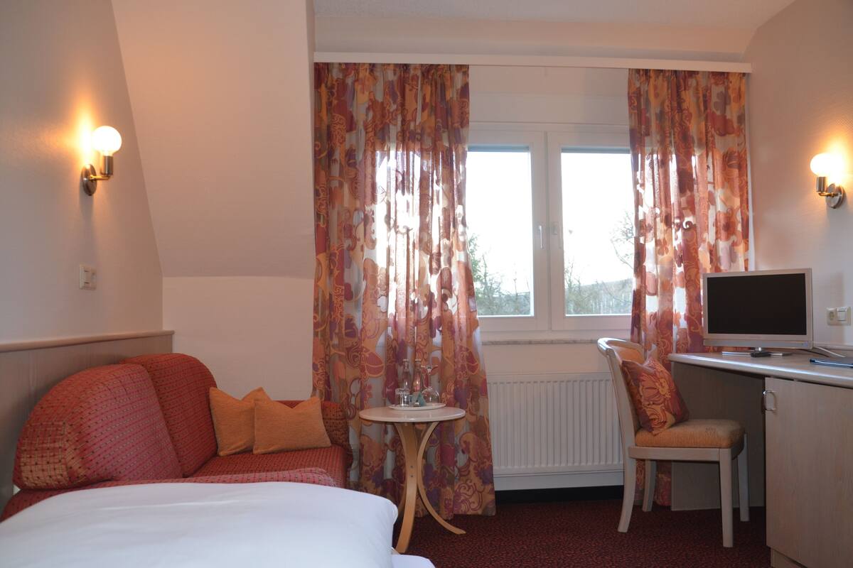 Ein Hotelzimmer mit einem Bett, einem Schreibtisch mit Computer, einem Stuhl und Vorhängen.