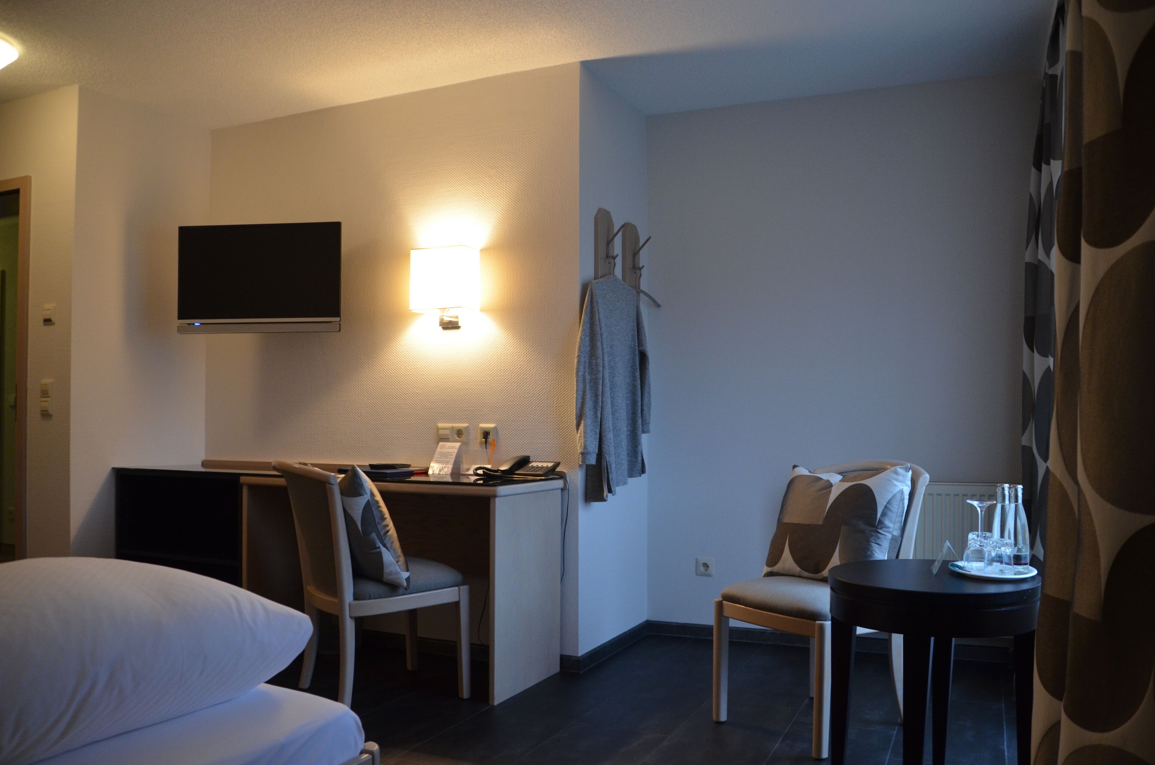 Habitación de hotel con cama, escritorio con silla, televisor montado en la pared y una mesita con vasos y una botella.