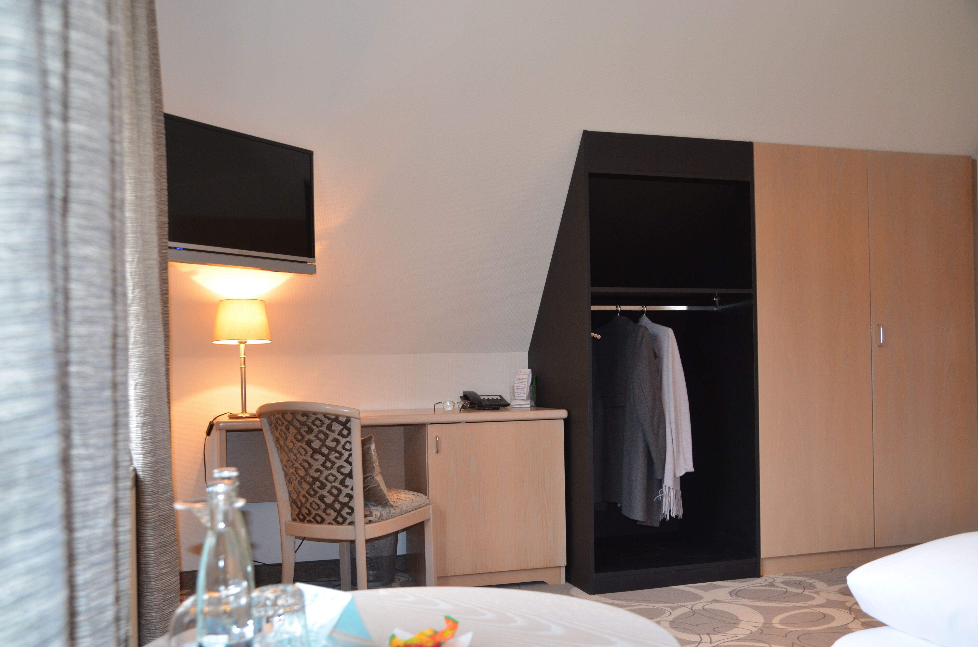 A hotel room with a desk, a chair, a TV mounted on the wall, a wardrobe, and a bedside table with a lamp.