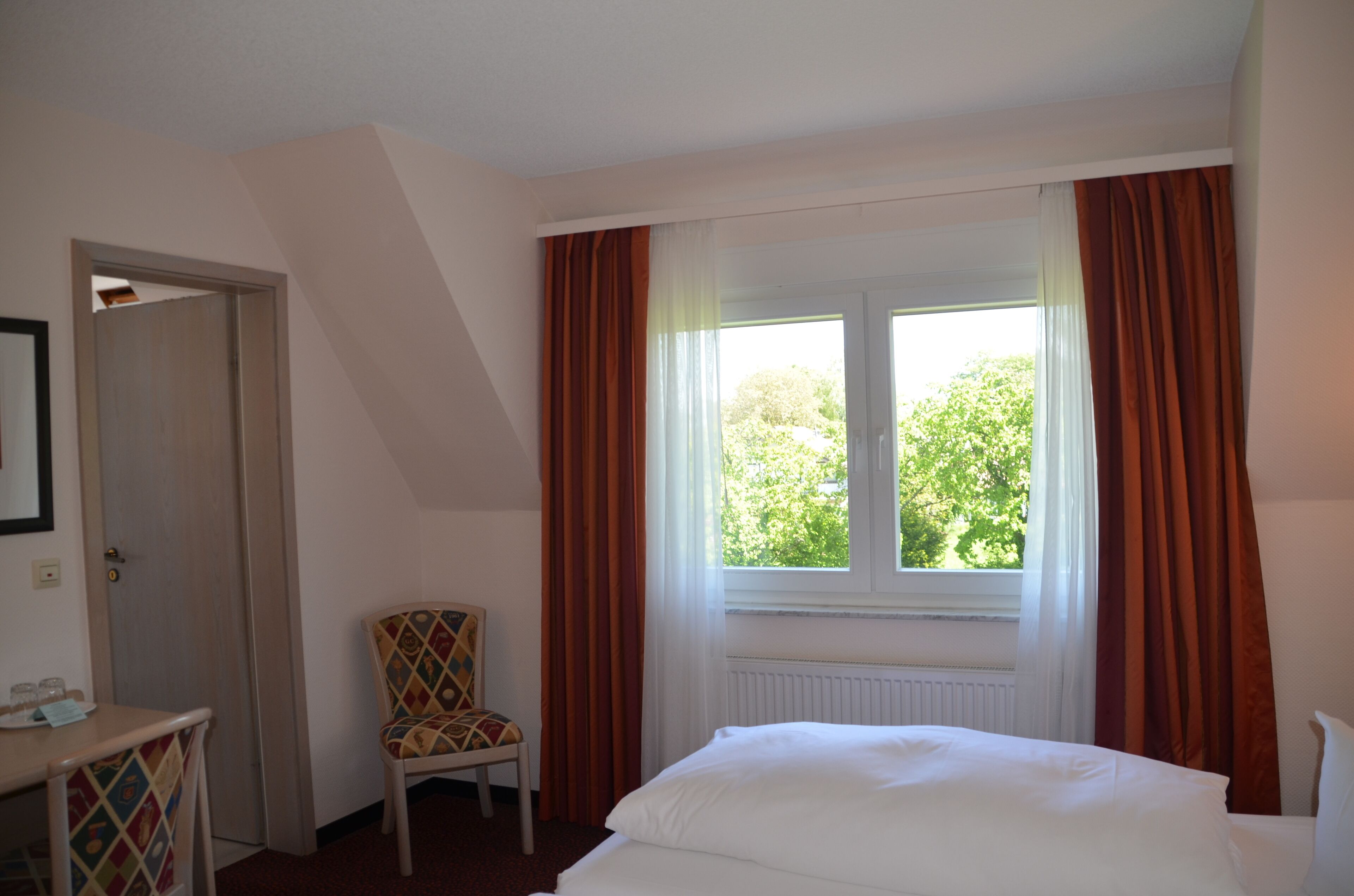 A hotel room with a bed, a chair, a table with glasses, and a window with curtains.