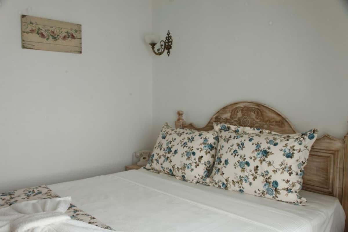 A bedroom with a wooden headboard, floral bedding, a bedside table with a lamp, and a wall-mounted light fixture.