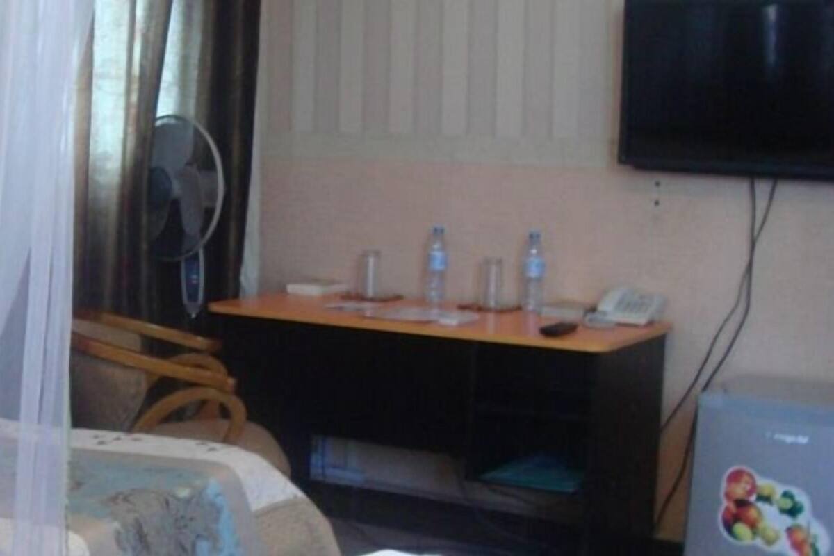 A hotel room with a bed covered by a mosquito net, a desk with a chair, a television mounted on the wall, and a small table with a phone and water bottles.