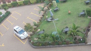 Vista aérea de um estacionamento com vários carros, uma área ajardinada com mesas ao ar livre e um caminho que leva à entrada de um edifício.