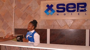 A hotel reception desk with a receptionist and a guest. The hotel name 'XSEB HOTEL' is visible on the wall. - Seb Hotel (Kampala)