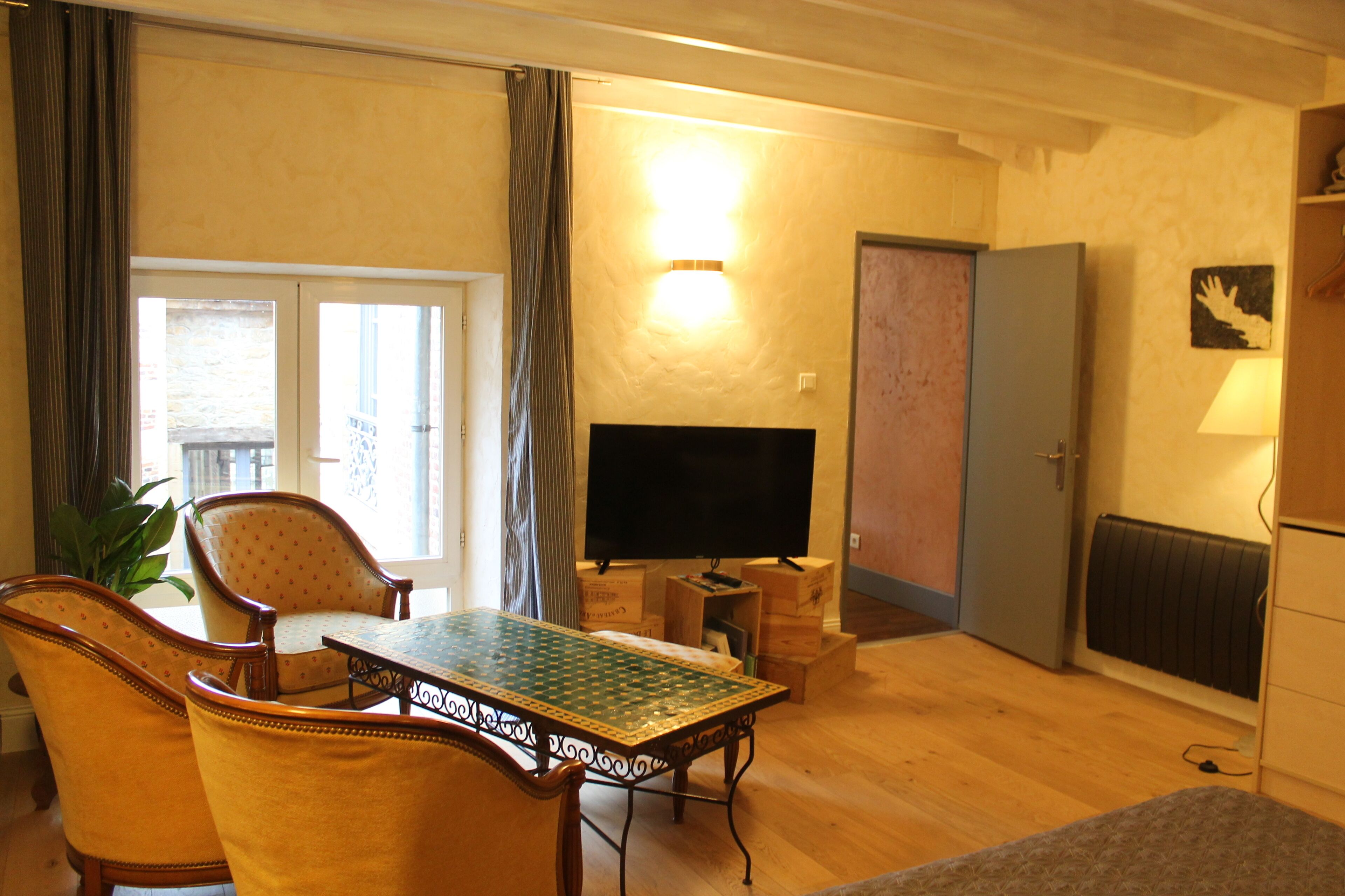 A living room with a wooden floor, a green patterned table, two yellow armchairs, a flat-screen TV, and a door leading to another room.