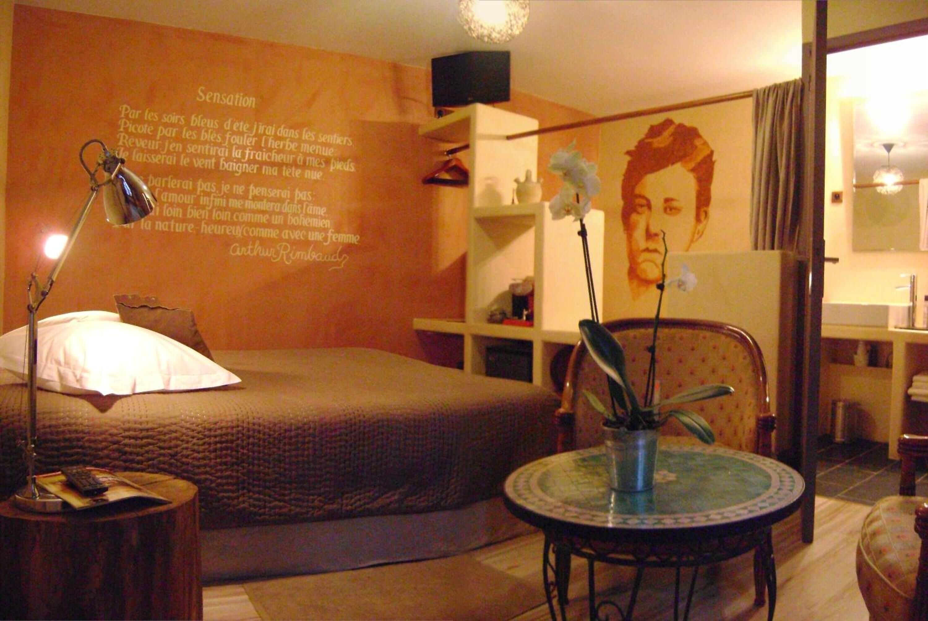 Un dormitorio con una cama, una lámpara de noche, una mesita con un florero y una pared decorada con una cita y un retrato.