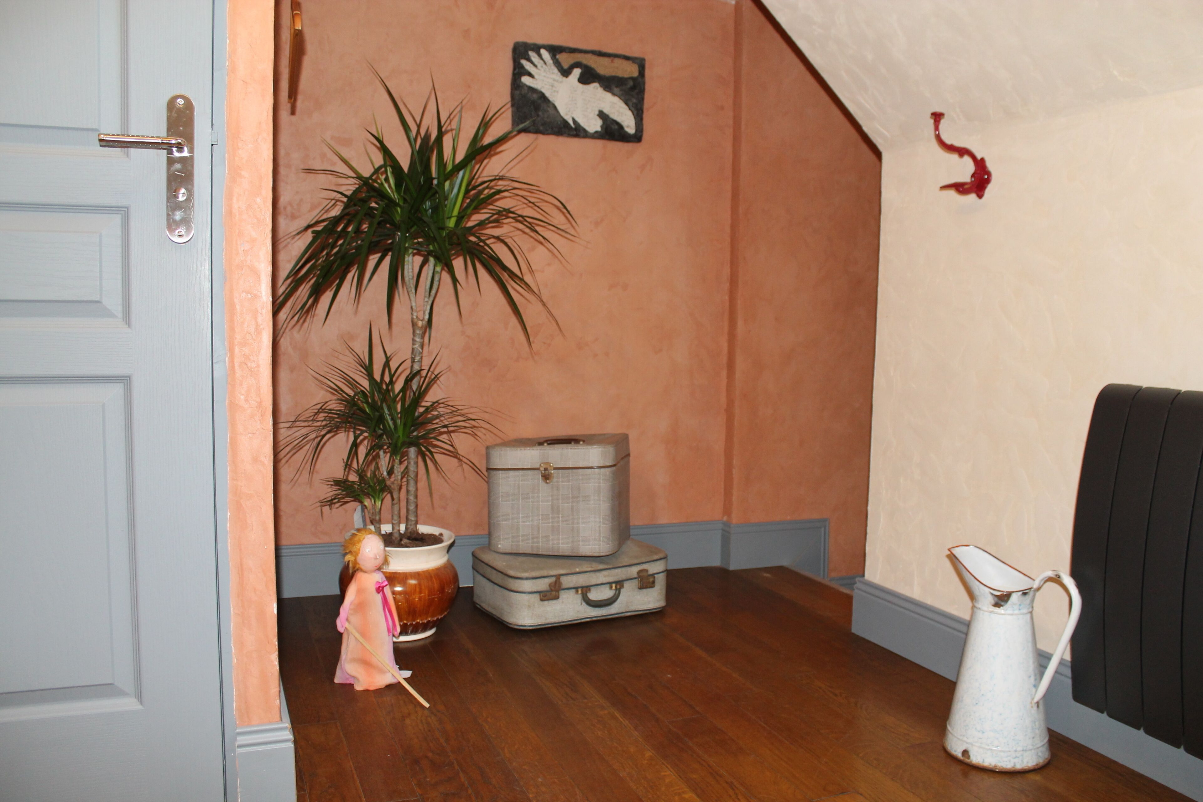 Ein Zimmer mit Holzboden, geschlossener Tür, Blumentopf, Puppe, einem alten Koffer und einer weißen Kanne.