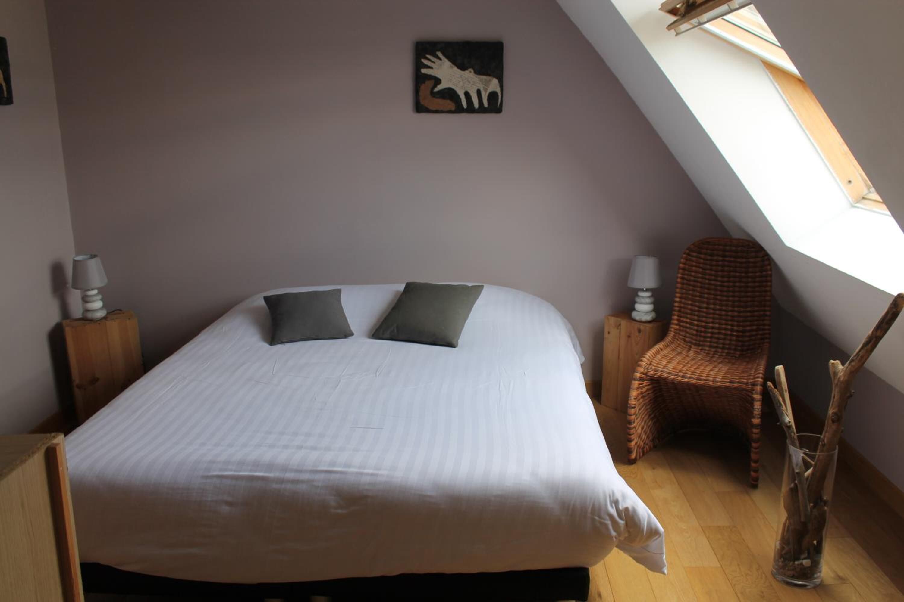 A bedroom with a large bed, two pillows, a wicker chair, a wall-mounted artwork, and a skylight.