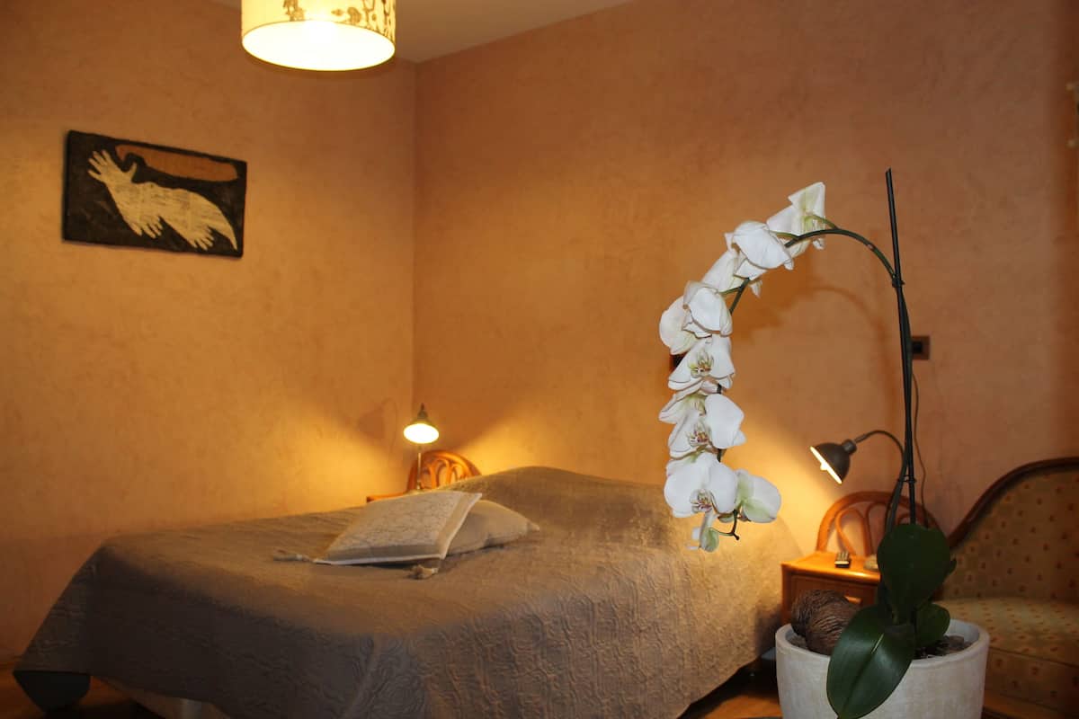 A bedroom with a bed, a nightstand, a lamp, a wall-mounted picture, and a potted orchid.