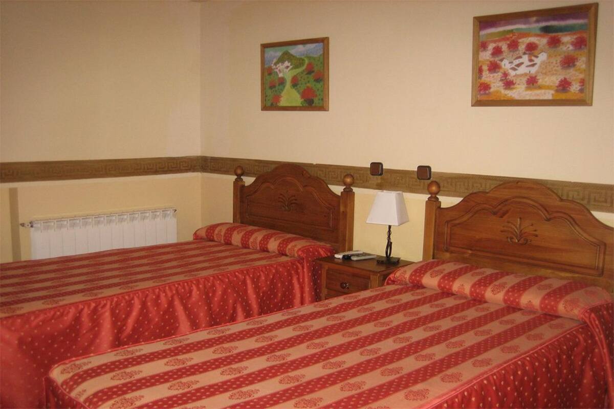 Une chambre d’hôtel avec deux lits, chacun recouvert d’une couvre-lit à motifs rouges et beiges. On y trouve une tête de lit en bois ornée d’un motif décoratif, une table de chevet avec une lampe et un petit tiroir. Les murs de la chambre sont de couleur beige et il y a une fenêtre avec un store blanc.