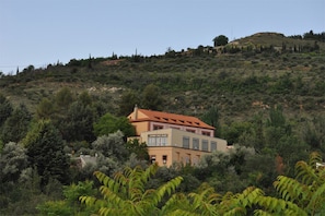 Um edifício de dois andares com telhado de telhas vermelhas, rodeado por vegetação densa e colinas ao fundo.