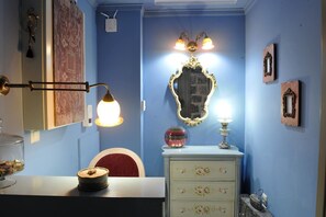 Ein Zimmer mit blauer Wand, einer weißen Kommode mit Blumenmotiven, einem Spiegel mit aufwendigem Rahmen und einer Lampe mit einer Kugelform-Leuchte.