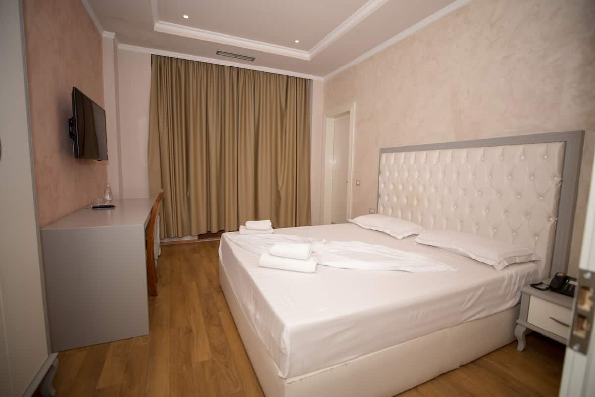 Habitación de hotel con una cama grande, una mesita de noche, un televisor montado en la pared y una ventana con cortinas.