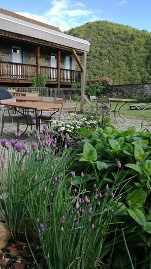 Un área de estar al aire libre con mesas y sillas, rodeada de plantas en flor y un edificio de madera con balcón.