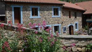 A stone building with a red-tiled roof, multiple windows with blue trim, and a stone wall with flowering plants in front. - Chambres Et Tables D'Hote De Margaridou (Blesle)