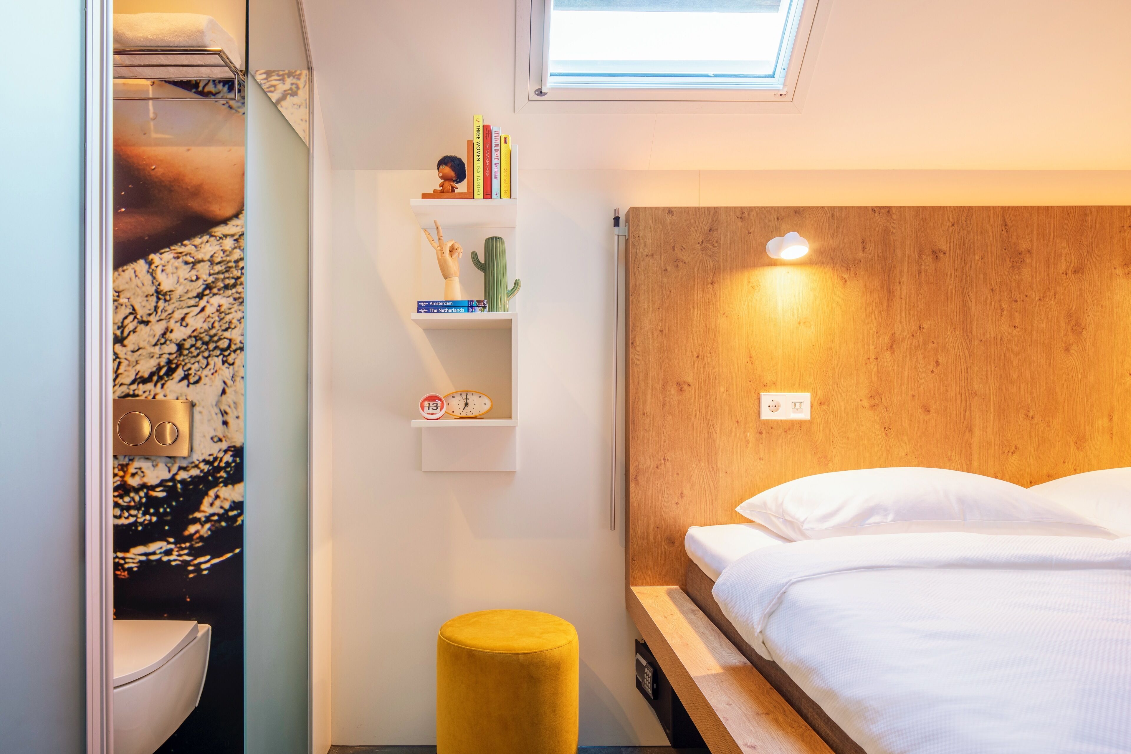 Un dormitorio moderno con cabecera de madera, una mesita de noche, un otoman amarillo y una estantería con libros y un cactus.