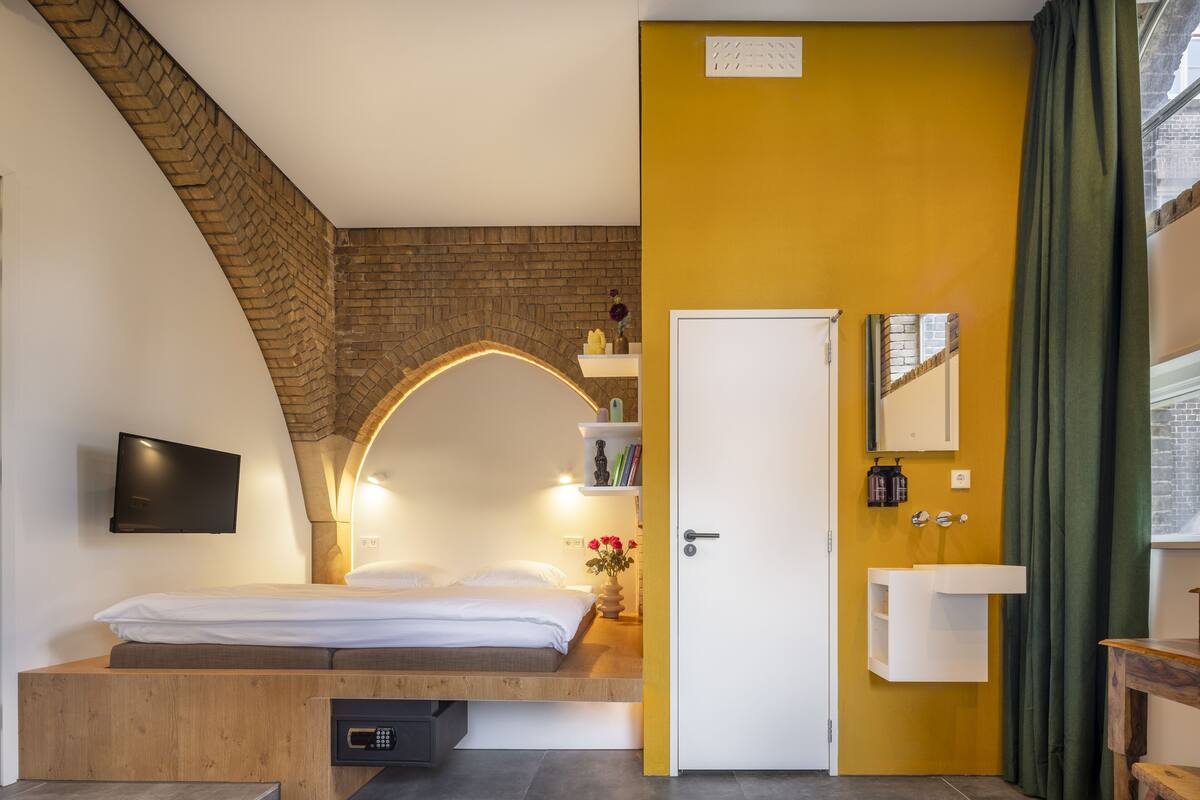 A modern bedroom with a bed, a TV, a built-in bench, a brick wall, a yellow accent wall, a bathroom with a sink and mirror, and a door.